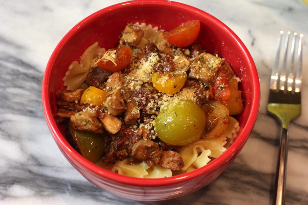 A red bowl filled with an easy slow cooker pesto chicken recipe served over bowtie pasta next to a silver fork on a white marble countertop.