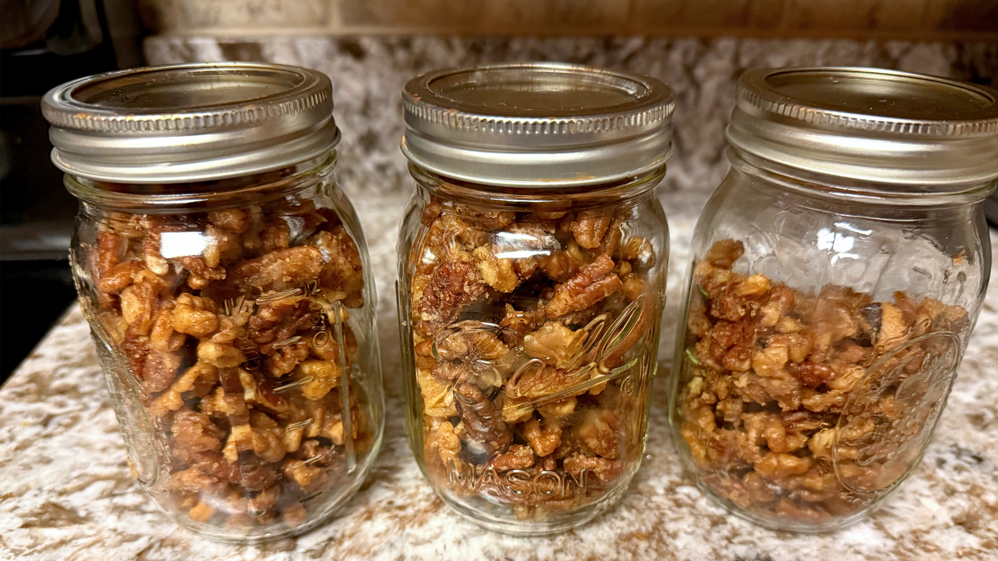 Two glass mason jars filled to the brim with glossy, roasted candied pecans and walnuts sitting on a light speckled kitchen countertop.