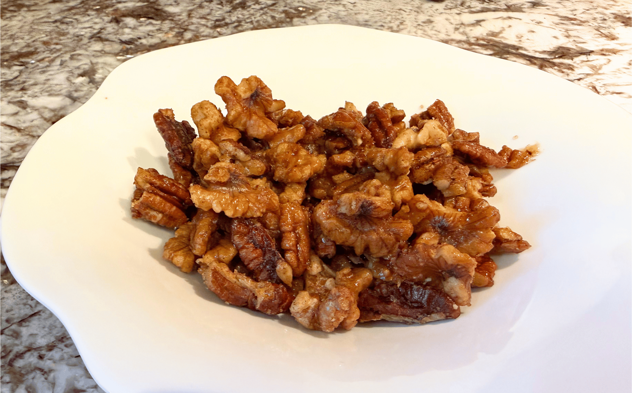 A close-up of a ceramic serving bowl overflowing with glossy, oven-roasted candied pecans and walnuts.