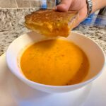A hand dipping a crispy, grilled cheese sandwich into a white bowl of creamy tomato soup on a wooden table.