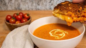 A hand dipping a crispy, gooey grilled cheese sandwich into a white bowl of creamy tomato soup on a wooden table.