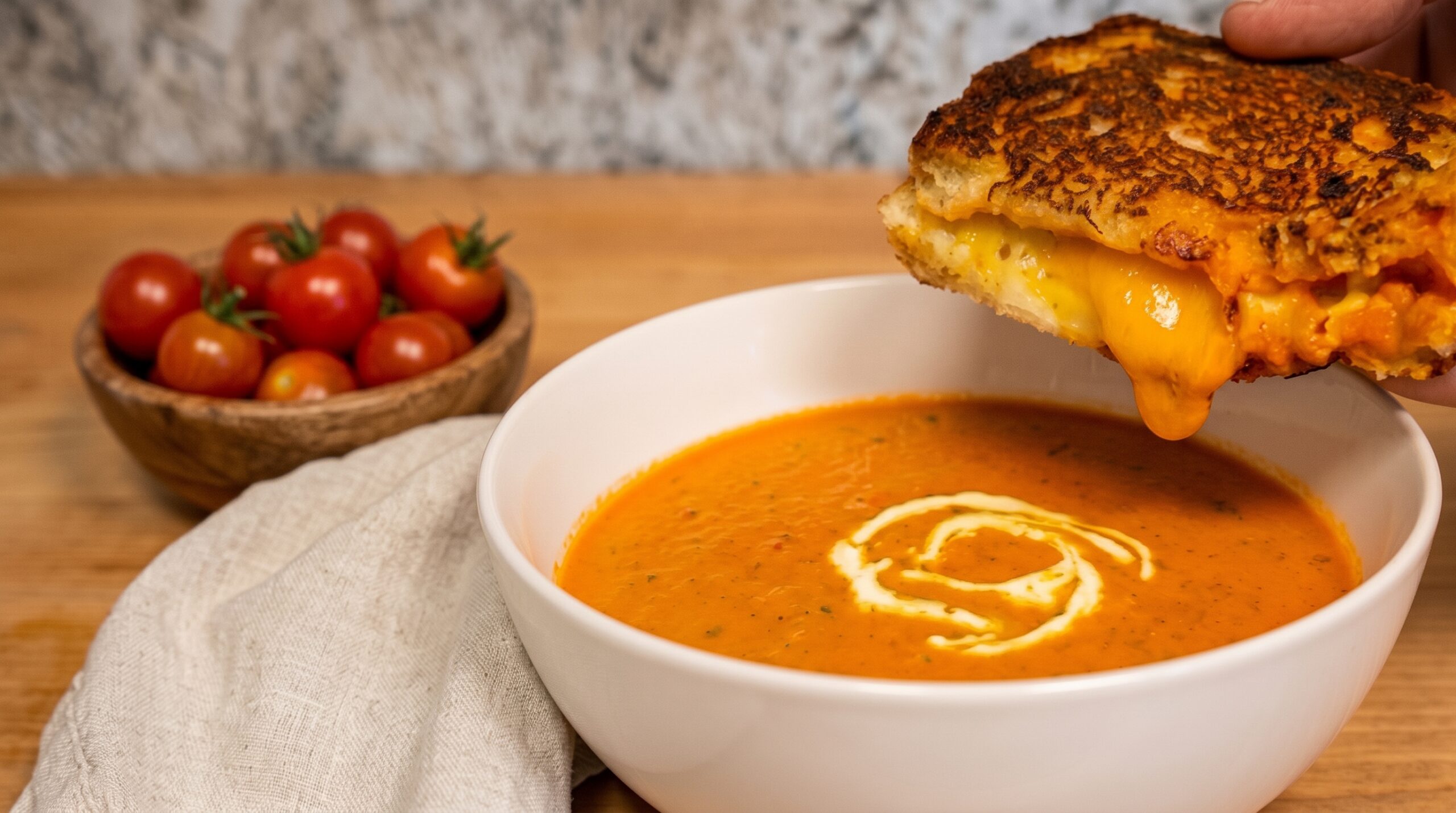 A hand dipping a crispy, gooey grilled cheese sandwich into a white bowl of creamy tomato soup on a wooden table.