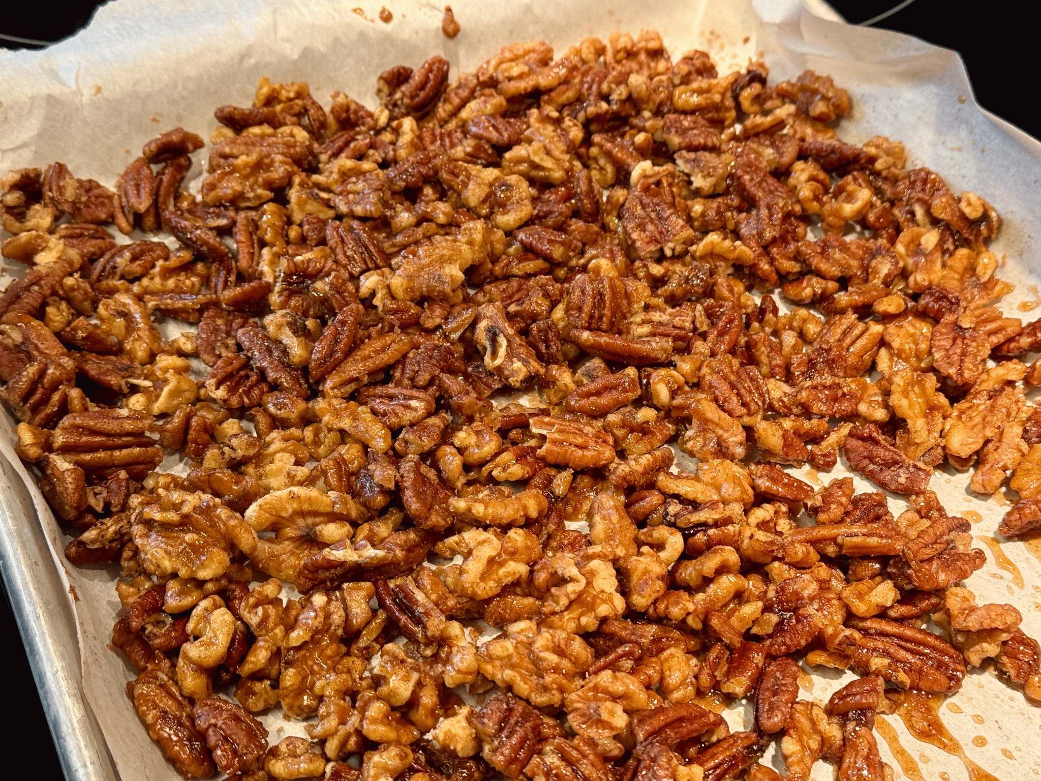Freshly baked candied spiced pecans and walnuts spread out in a single layer on a parchment-lined metal baking sheet to cool.